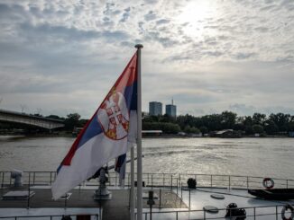 Serbian flag on a boat with an urban skyline and river scene in daytime.