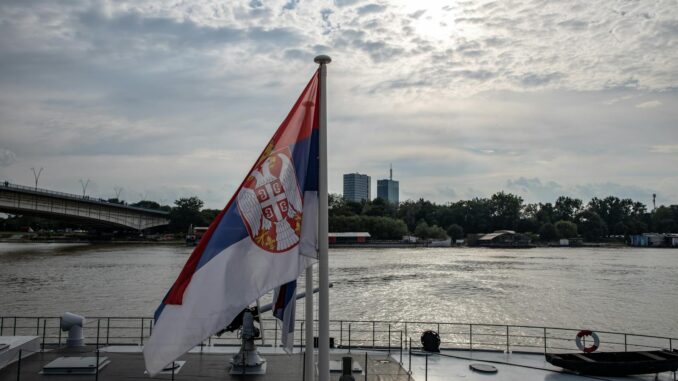 Serbian flag on a boat with an urban skyline and river scene in daytime.