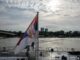 Serbian flag on a boat with an urban skyline and river scene in daytime.